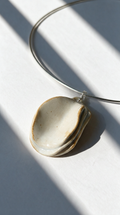 A minimalist silver wire hoop necklace with a small, organic-shaped cream-colored ceramic pendant, styled on a light gray surface with dramatic diagonal window shadows.
