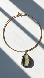 A gold-plated wire hoop necklace with a teardrop-shaped green stone pendant, displayed on a white surface with diagonal window shadows.