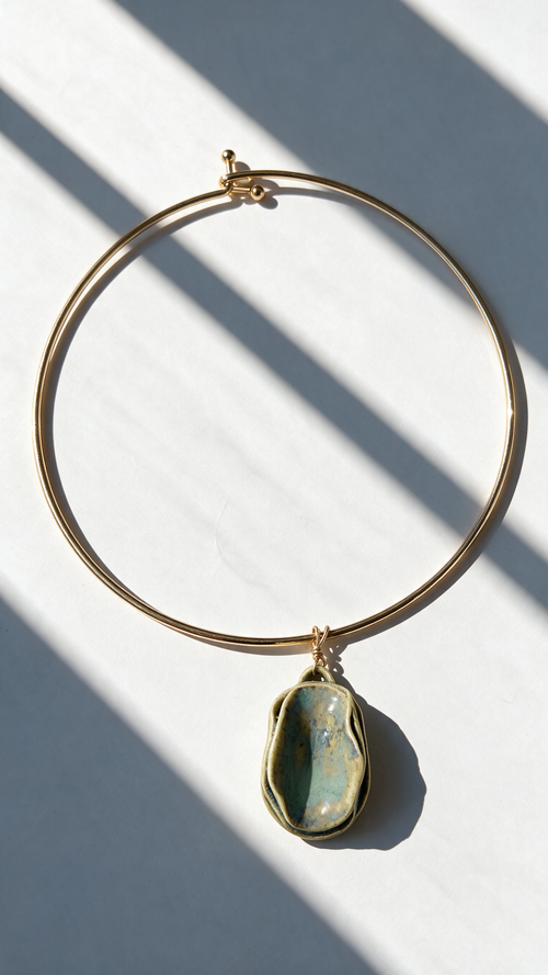 A gold-plated wire hoop necklace with a teardrop-shaped green stone pendant, displayed on a white surface with diagonal window shadows.