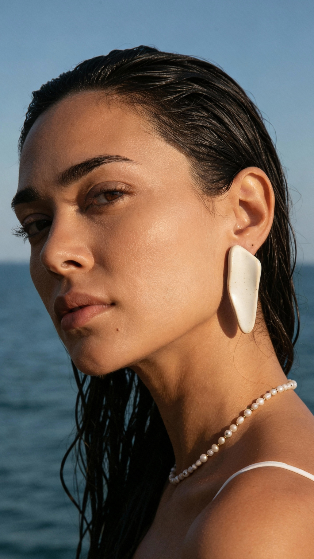 Woman wearing Galini white ceramic statement earrings on the beach, model with wet hair against the sea