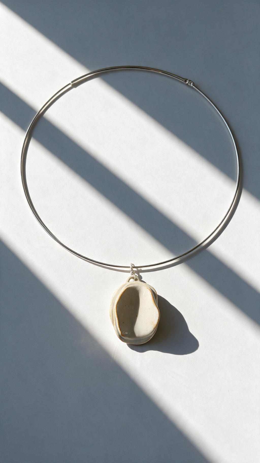 A minimalist silver wire hoop necklace with a small, organic-shaped cream-colored ceramic pendant, styled on a light gray surface with dramatic diagonal window shadows.