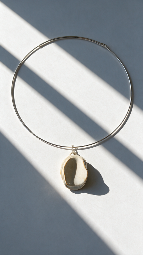 A minimalist silver wire hoop necklace with a small, organic-shaped cream-colored ceramic pendant, styled on a light gray surface with dramatic diagonal window shadows.