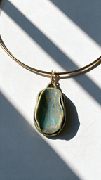 A gold-plated bangle necklace with a large, irregularly shaped greeglazed pendant in shades of teal, blue, and gold, resting on a white surface with diagonal shadows. 