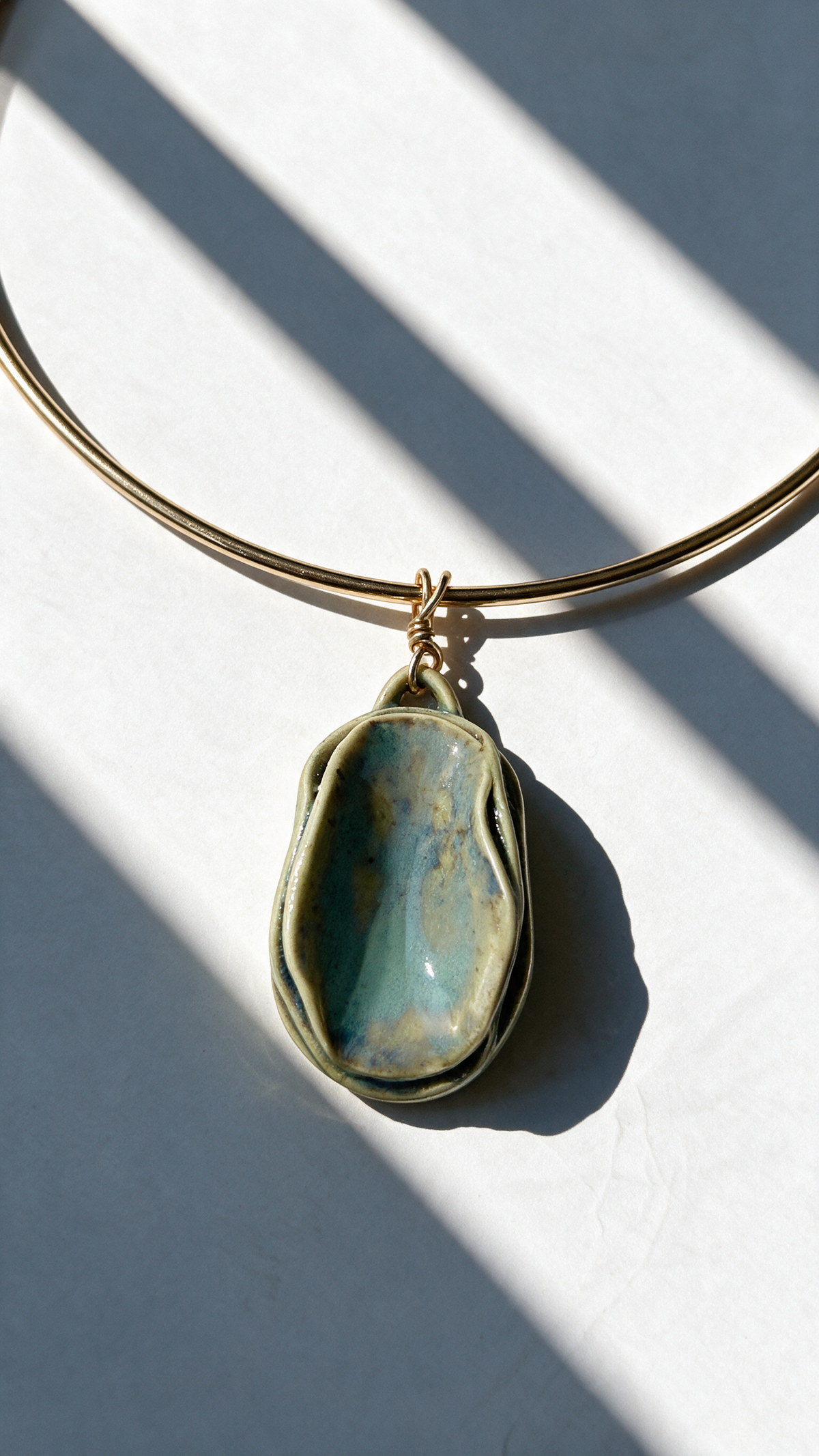 A gold-plated bangle necklace with a large, irregularly shaped greeglazed pendant in shades of teal, blue, and gold, resting on a white surface with diagonal shadows. 
