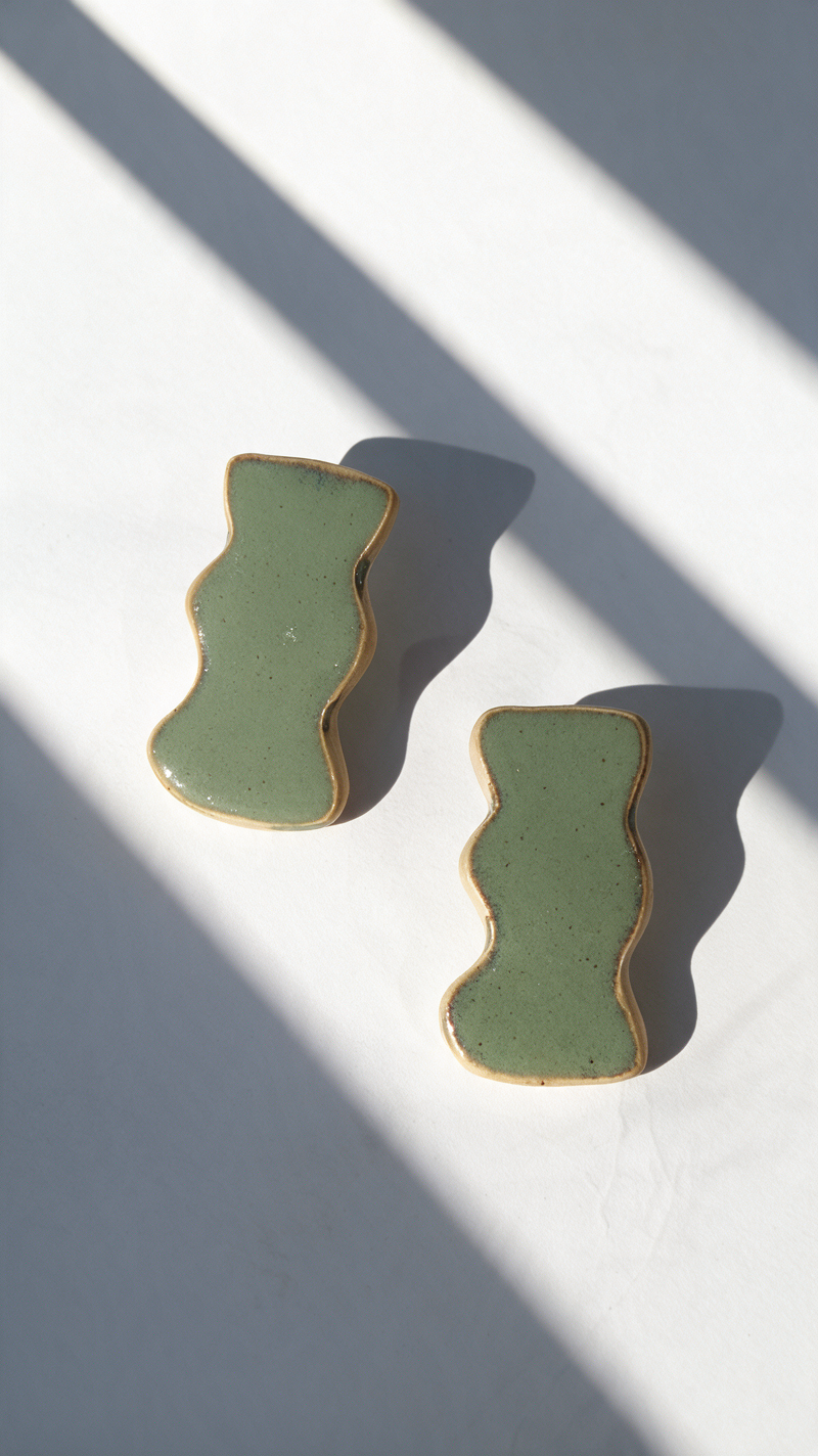 Two handmade ceramic jewelry with white olive green glaze and speckles, displayed diagonally on a white surface with dramatic window shadow patterns.