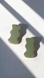 Two handmade ceramic jewelry with white olive green glaze and speckles, displayed diagonally on a white surface with dramatic window shadow patterns.