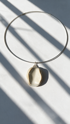 A minimalist silver wire hoop necklace with a small, organic-shaped cream-colored ceramic pendant, styled on a light gray surface with dramatic diagonal window shadows.