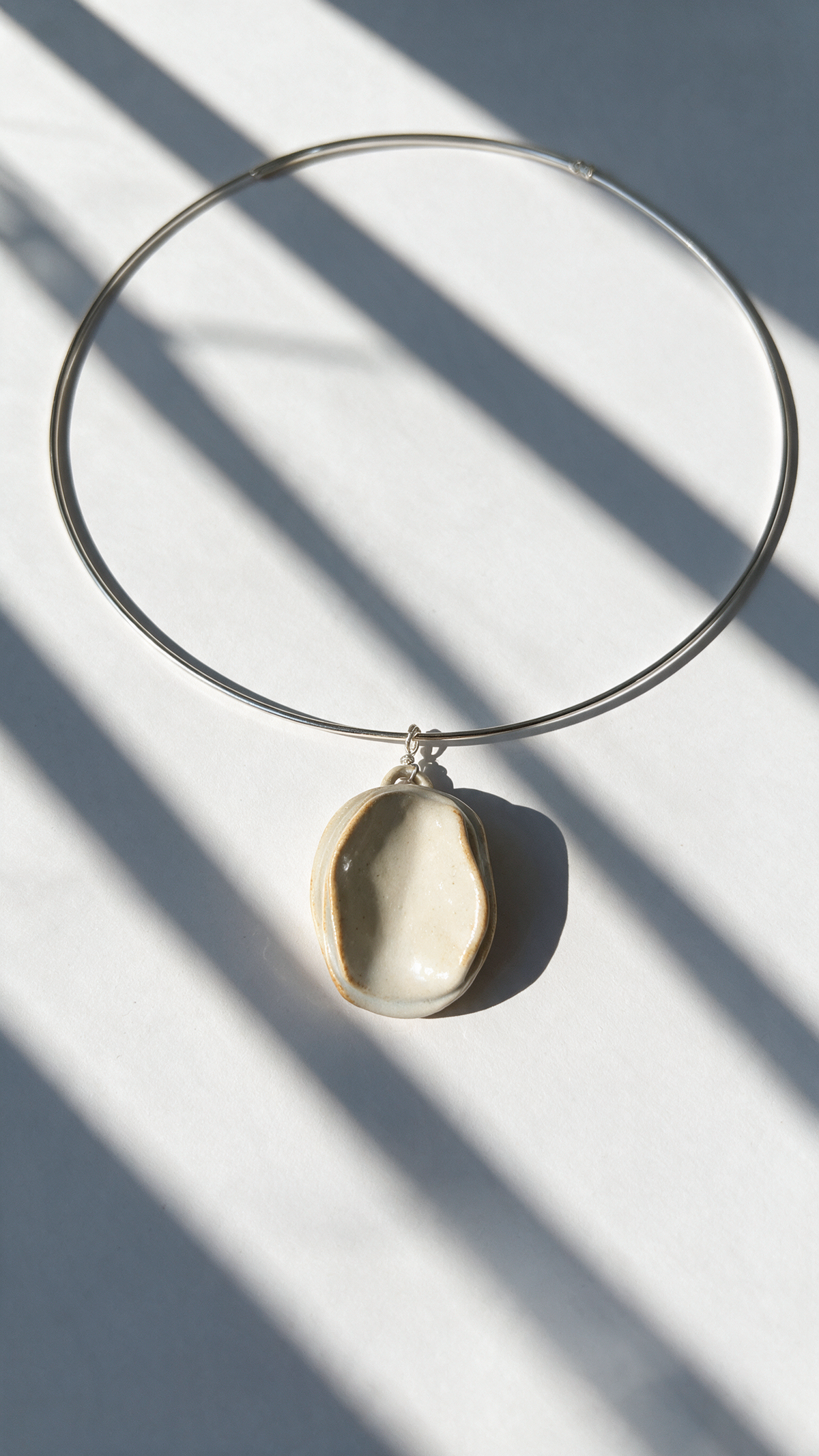 A minimalist silver wire hoop necklace with a small, organic-shaped cream-colored ceramic pendant, styled on a light gray surface with dramatic diagonal window shadows.